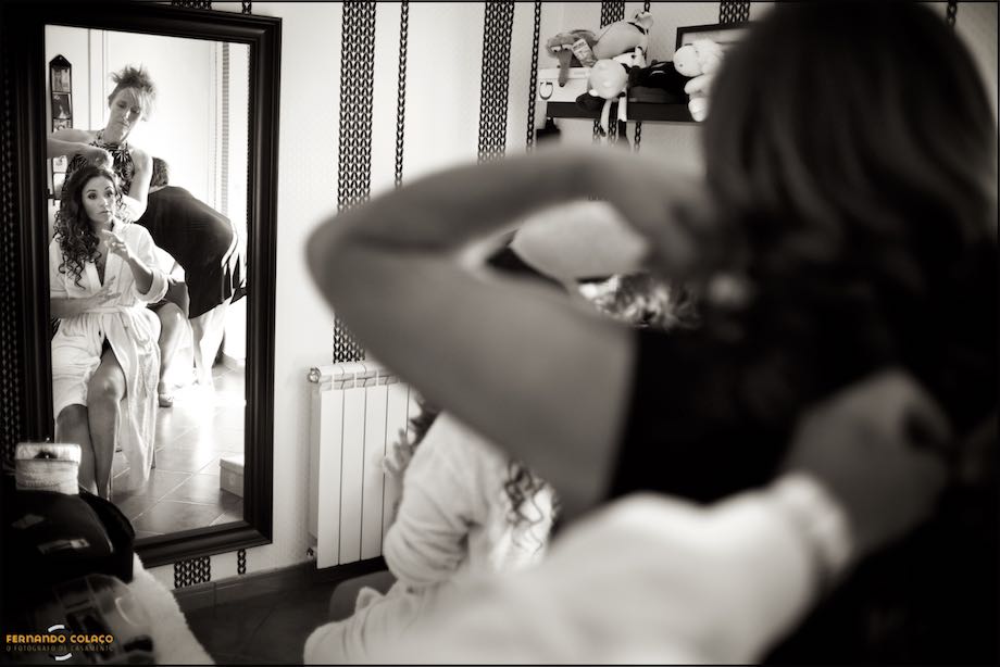 Num espelho, no lado esquerdo, a noiva sendo penteada com a mão alguém que abotoa um vestido por detrás, desfocados, do lado direito.