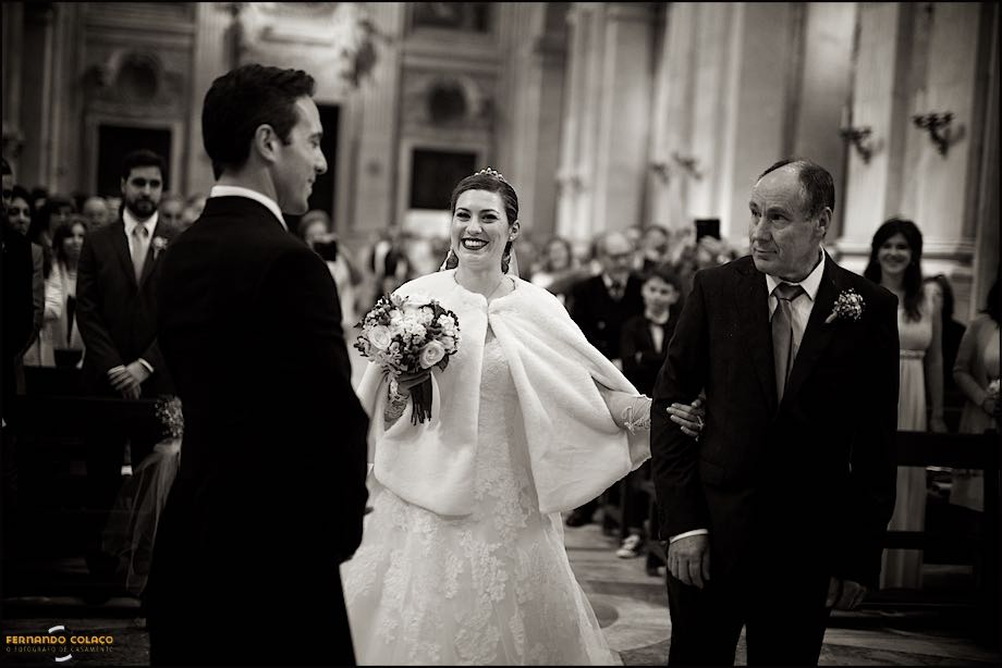 Noiva, sorridente, com o pai chegam em frente do noivo, no altar para a cerimónia do casamento.