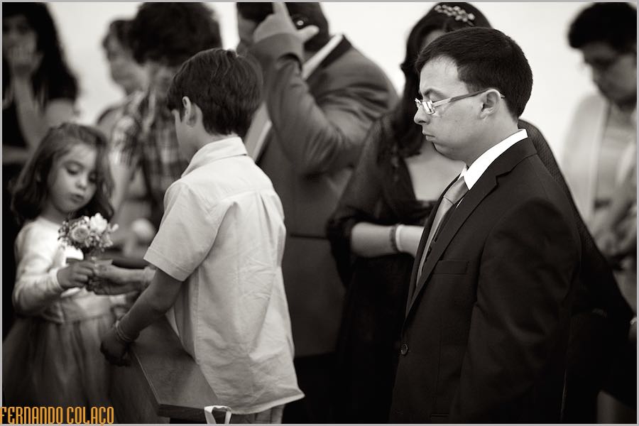 Um convidado do casamento na cerimónia, jovem e entediado perto de uma menina que entrega um ramo de flores a um rapazinho, vistos pelo fotógrafo de casamento no Alentejo.