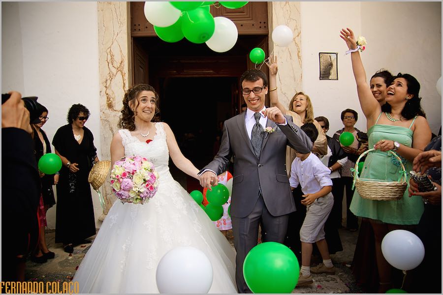 O noivo e a noiva saem na porta da igreja, depois da cerimónia por entre balões brancos e verdes e saudados pelos convidados.