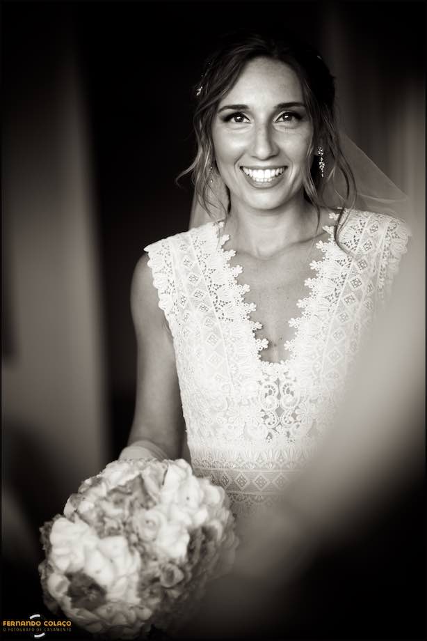 Já pronta para ir para ir para a cerimónia do casamento na Basílica de Mafra, a noiva sorri, entre desfocados, com o bouquet na mão.