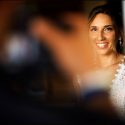 A noiva, pronta para partir para a cerimónia do casamento, sorri para alguém, muito desfocado, que lhe tira uma fotografia, vista pelo fotógrafo de casamento.