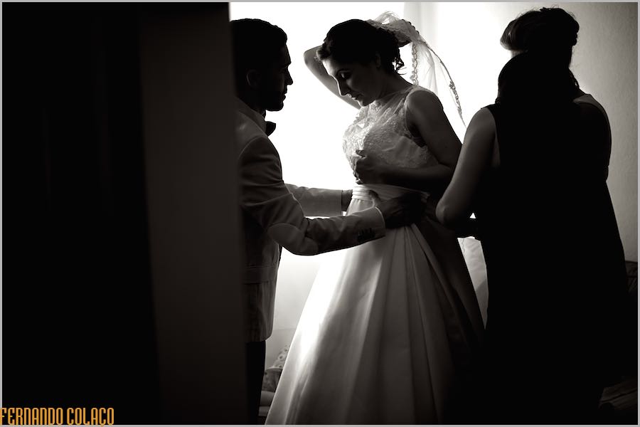 A noiva, entre a mãe e um a amigo que a ajudam a ajustar o vestido para a cerimónia do casamento.