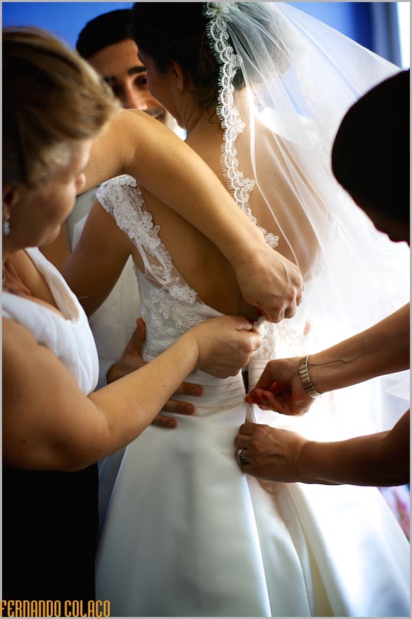 Quatro mãos apertam o vestido da noiva por detrás, vistos pelo fotógrafo de casamento.