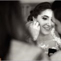 Emoldurada entre dois rostos desfocados, a face da noiva sorridente enquanto lhe põem os brincos, vista pelo fotógrafo de casamento.