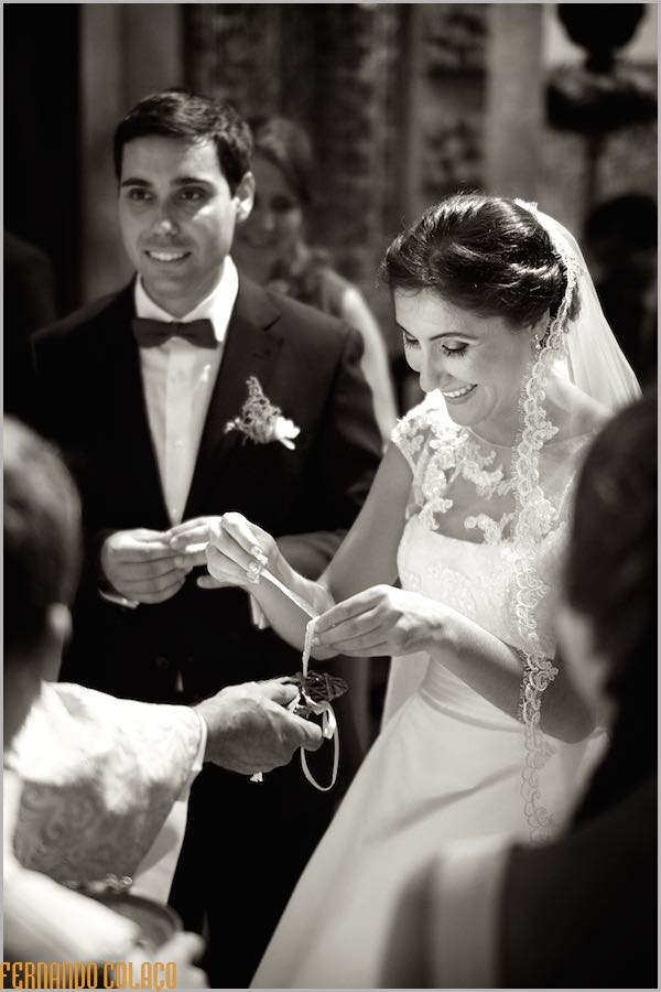 O noiva sorrindo, ao lado do noivo, retira as alianças do arranjo, que as trouxe até ali, para as colocar nos dedos e finalizar a cerimónia do casamento.