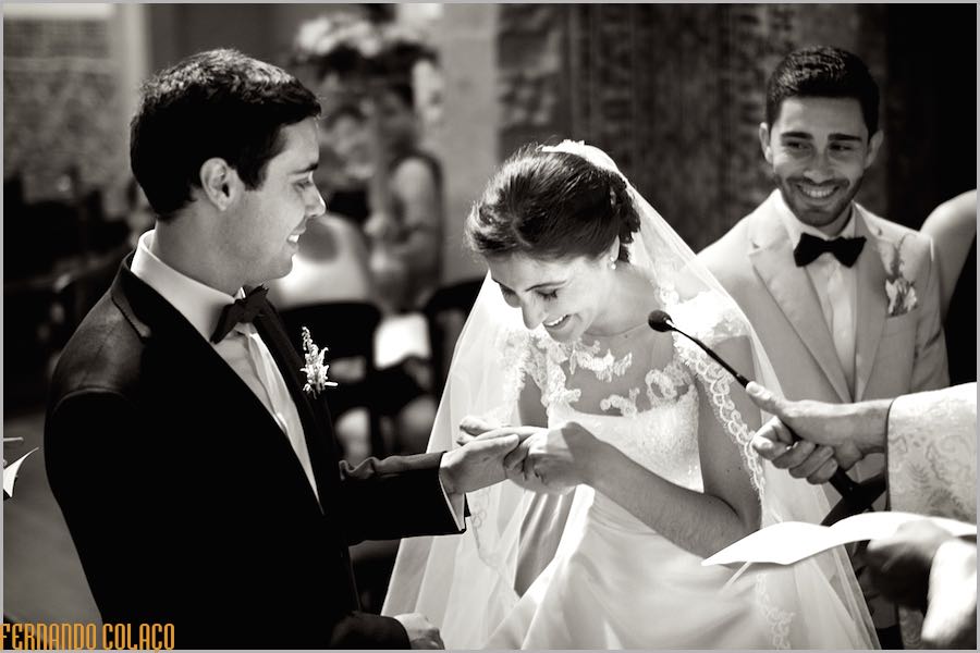 Rindo de satisfação a noiva entrega a aliança ao noivo, à sua frente, na cerimónia do casamento.