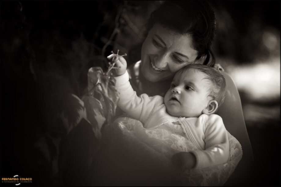 A smiling mother with her baby daughter in her arms, when she plays with a flower, in a composition by the christening photographer in Lisbon.