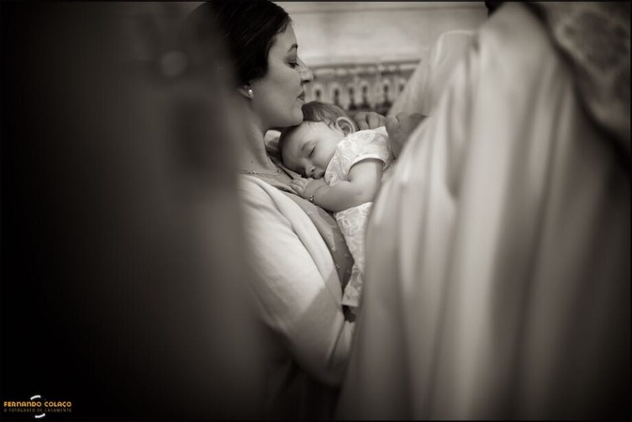 Just before the ceremony at the baptismal font, the baby sleeps on her mother's lap, seen among other family members, out of focus.