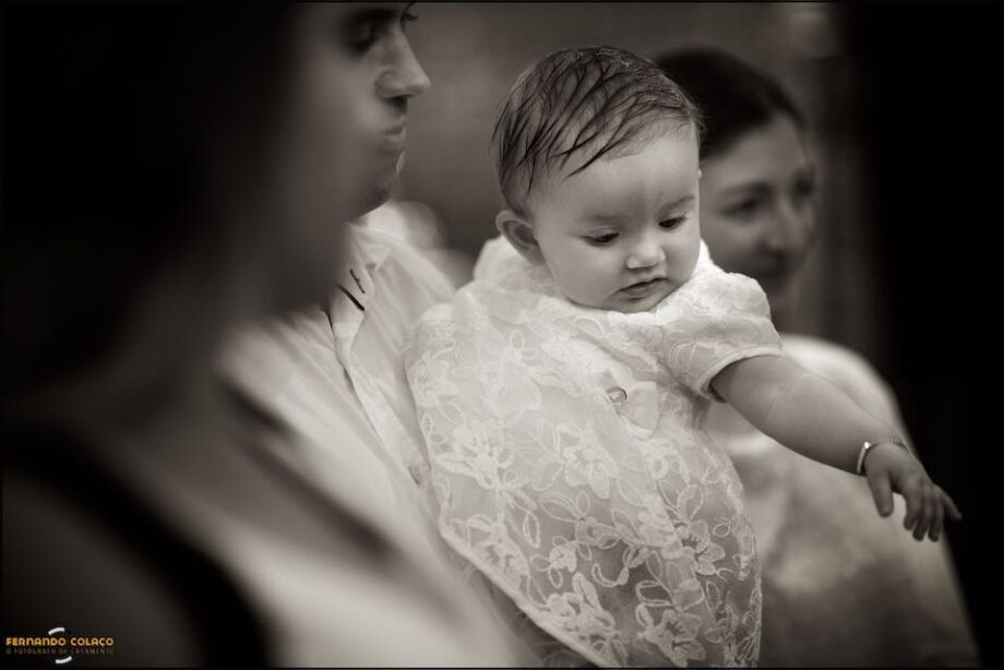 On her father's lap, at another moment of the baptism ceremony, the baby girl points to something looking down.