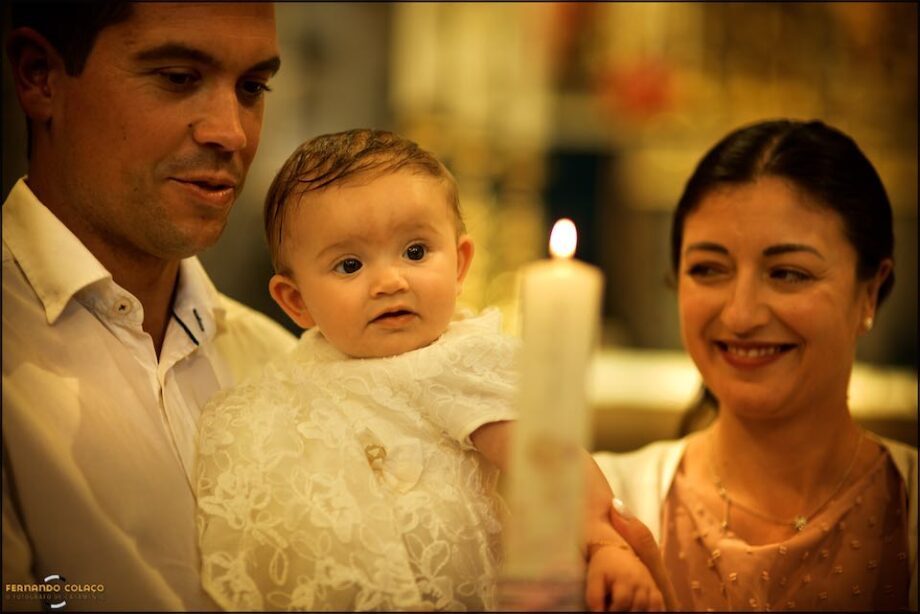 On her father's lap and next to her mother, during the baptismal candle ceremony, the baby girl looks, as if enchanted, at the light emanating from the candle.
