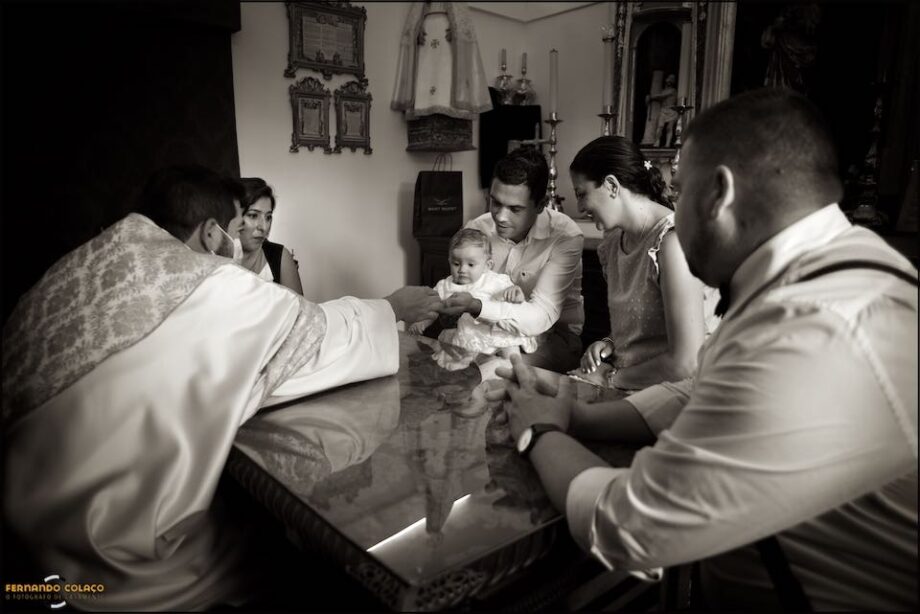 Sitting at the table for the baptismal signatures, the parents, the godparents and the priest all look at the newly baptized girl.