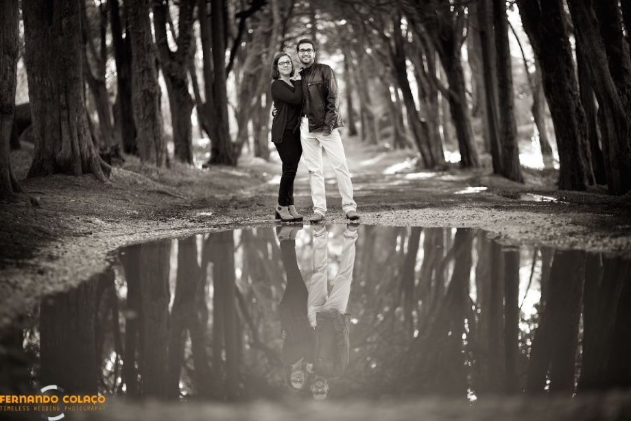 Depois de uma chuvada, o casal numa sessão de pré casamento, numa estrada ladeada de grandes árvores, na Peninha em Sintra, em frente de uma grande poça de água com o seu reflexo, vistos pelo fotógrafo de casamento em Lisboa.