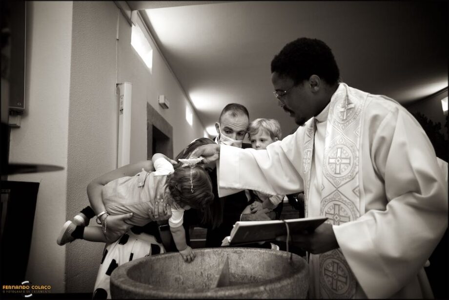 O momento em que o padre derrama a água baptismal na cabeça do menino a ser baptizado, segurado pela mãe e visto pelo pai e pelo irmão.