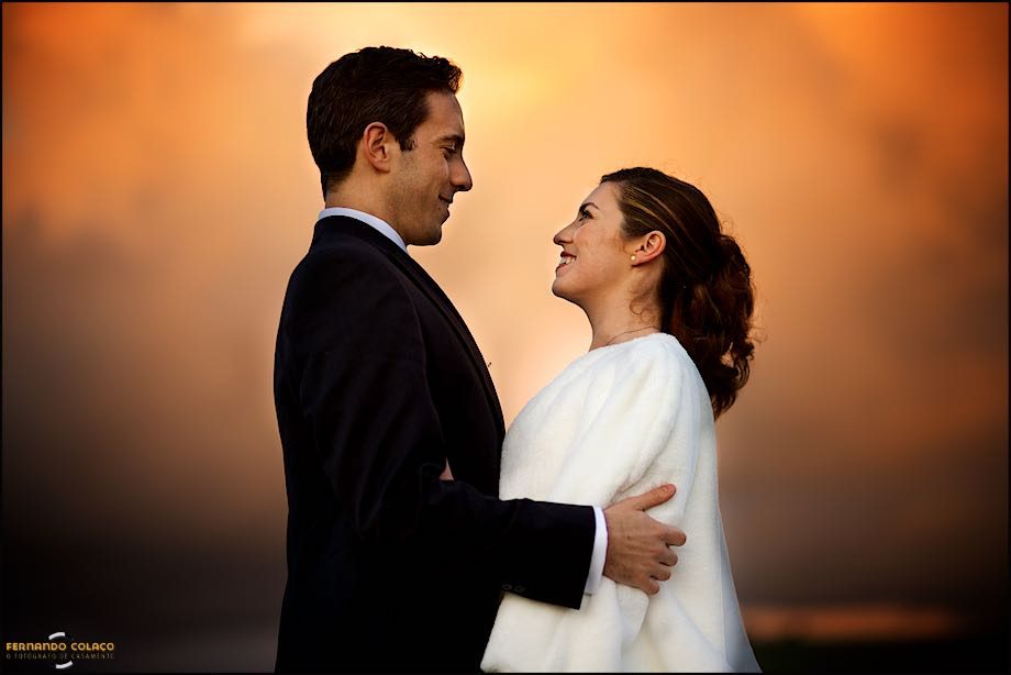 Fotografia a cores do noivo e da noiva a olharem um para o outro, com um céu nublado muito alaranjado por detrás.