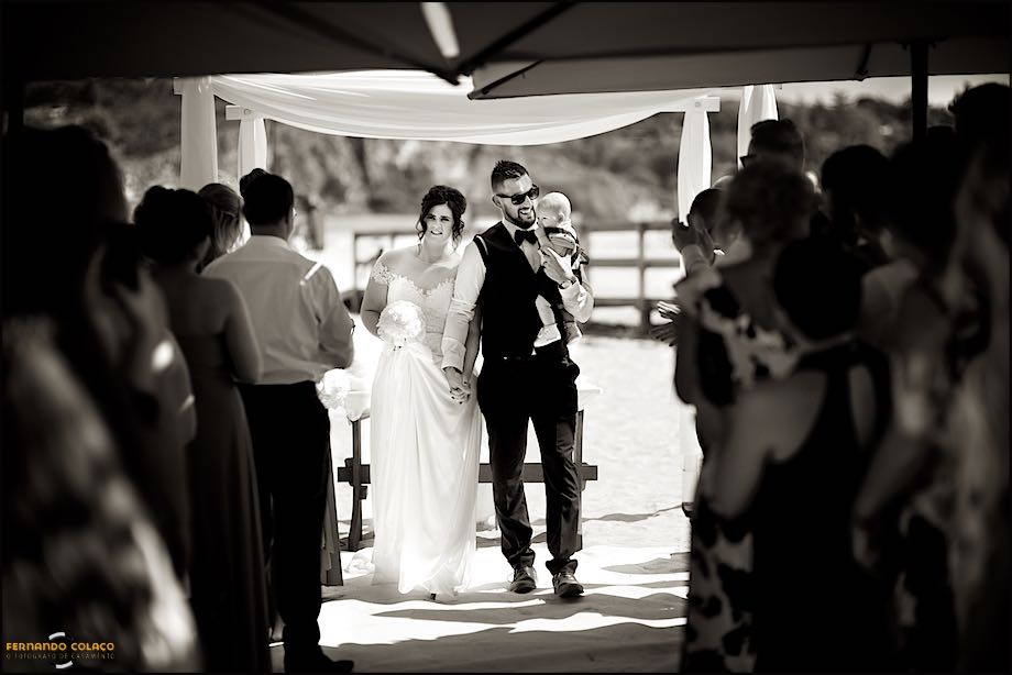 Noivos com o filho bebé, ao colo, saindo do local da cerimónia, no Club Nau, por entre os convidados, pelo fotógrafo de casamento no Algarve.