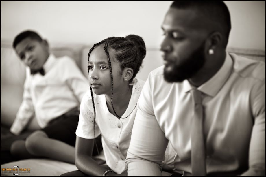 Rapariga, sentada, entre adulto e outra criança na preparação do noivo.