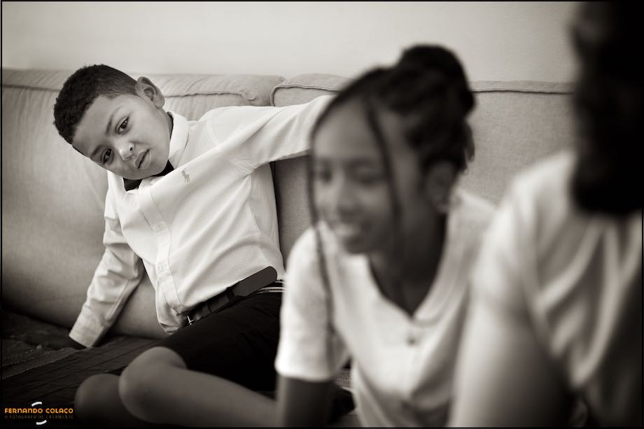 Filho do noivo, sentado num sofá, com dois convidados desfocados.