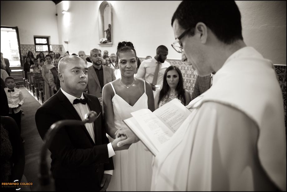 Noivos ouvindo o padre, à sua frente, para a cerimónia das alianças vistos pelo fotógrafo de casamento.