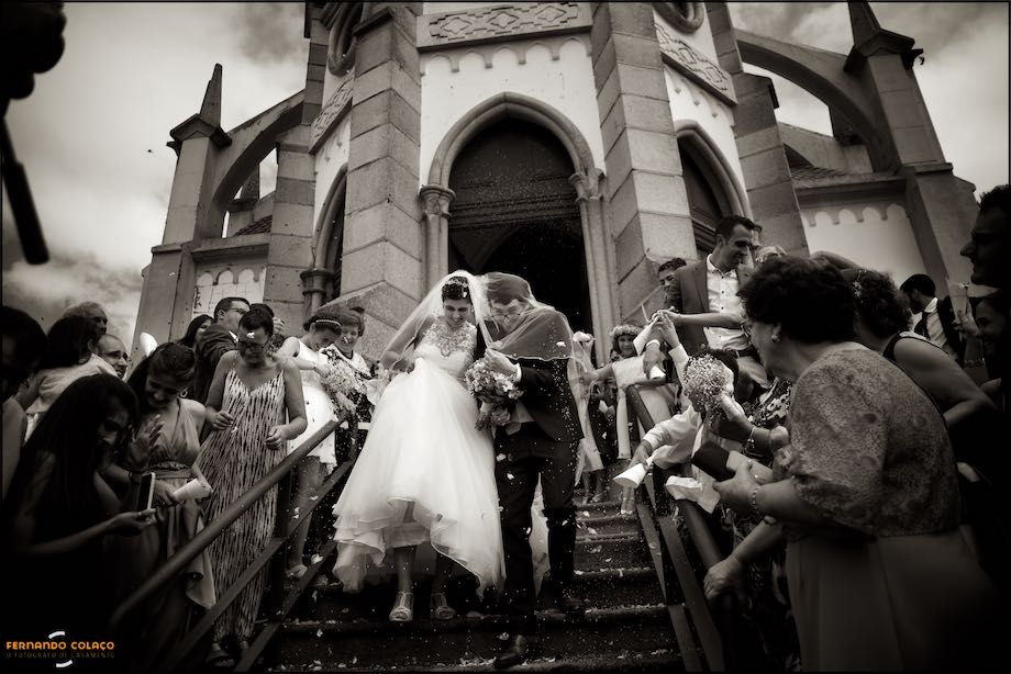 Noivos, convidados e igreja, festeja o casal de recém casados que desce a escadaria.