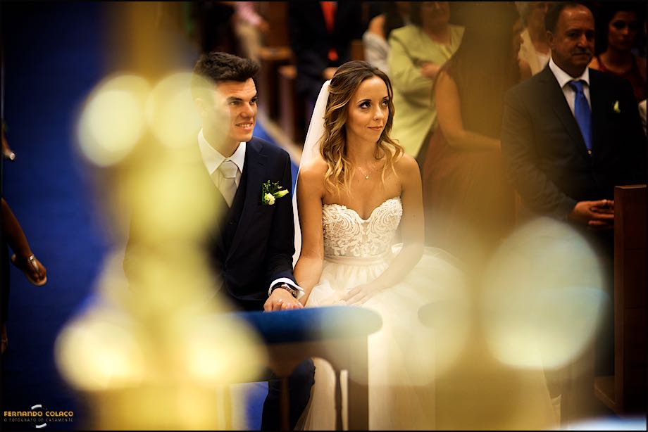 Por entre luzes desfocados, os noivos sentados em frente ao altar.