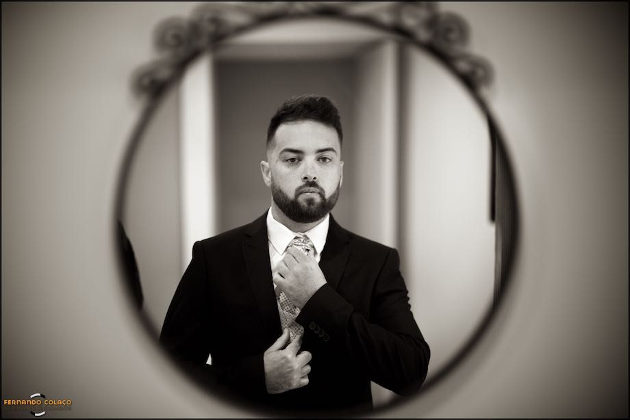 Groom in the mirror putting the finishing touches on his tie, in a composition of the wedding photographer.