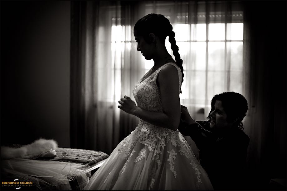 Silhouette of the bride and her sister helping her to button her wedding dress, in a composition of the wedding photographer in Portugal.