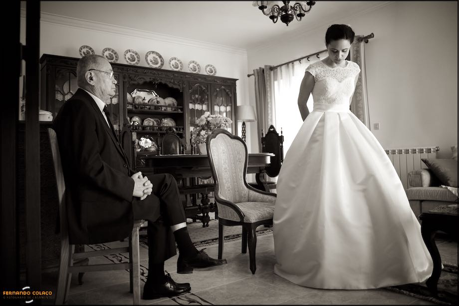 Father of the bride, seated in a chair, and bride standing showing him the dress, by the wedding photographer in Cascais/Estoril.