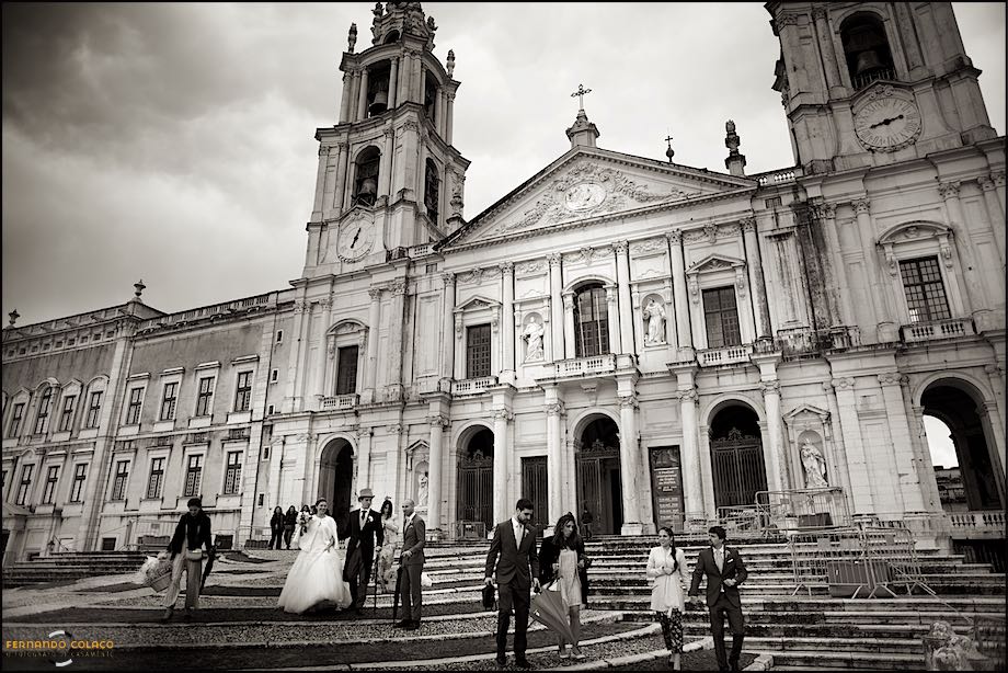 Bride and groom and guests coming down the stairs with the Basilica of Mafra in the back.