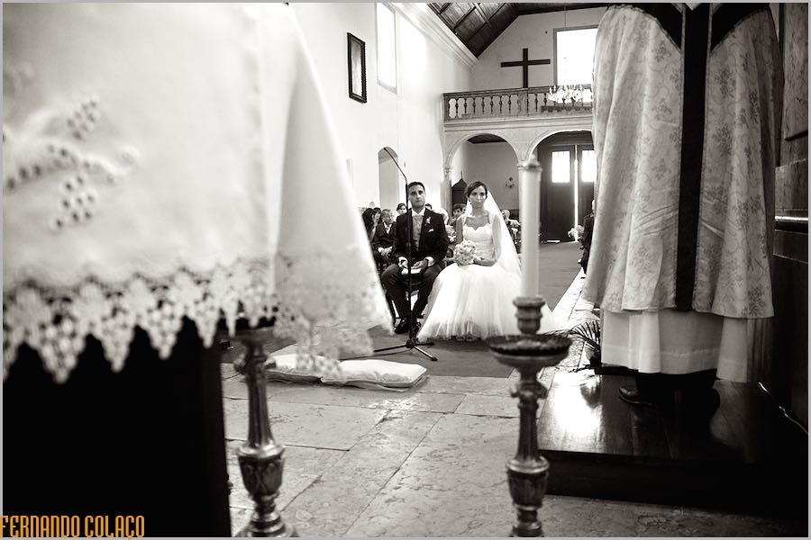 Between the altar table and the priest's robes, the bride and groom, seated, listen to the priest during the ceremony.