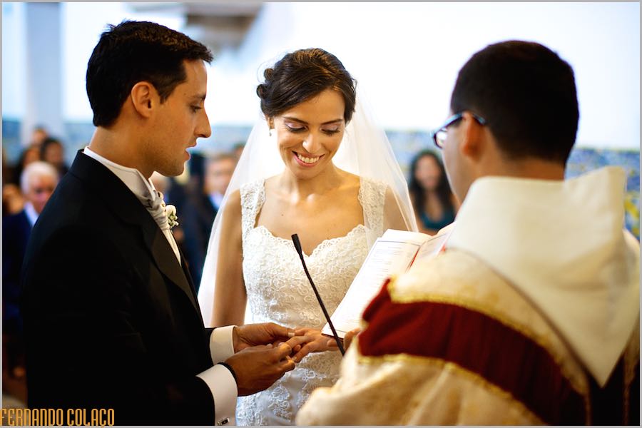 The bride, smiling, receives the ring given by the groom, together with the priest.