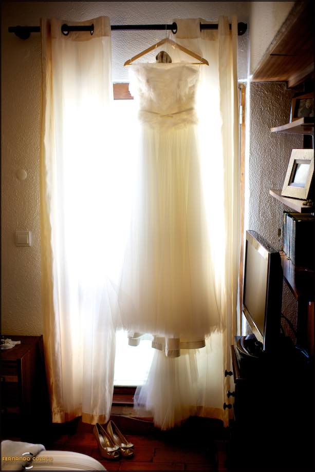 The dress of the bride hanging on the window, near the shoes.