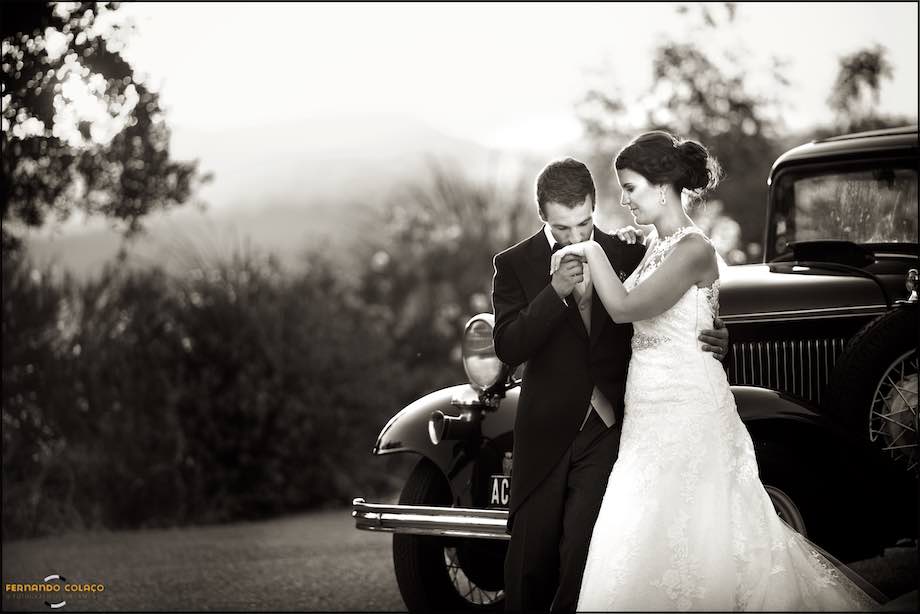The groom kisses the bride's hand, next to an old car, in the garden of Casa de Reguengos where the wedding party takes place.
