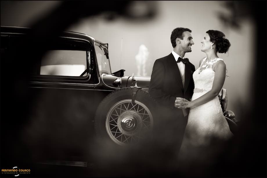 As in an old movie, the bride and groom laugh facing each other and holding hands, leaning against a vintage car, from the time of these films.