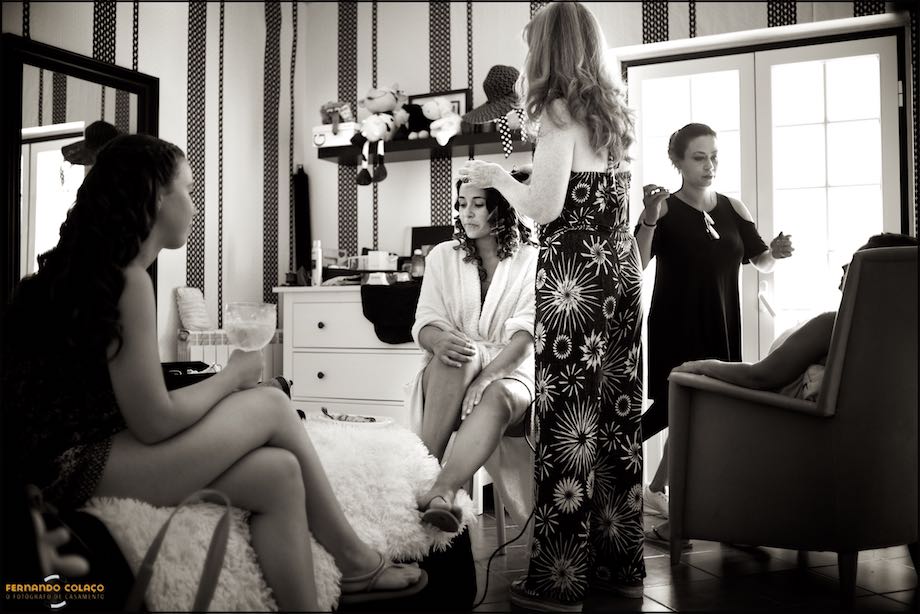 The bride, seated, between a hairdresser, a make-up artist and two wedding guests, as they take care of her hairstyle for her wedding day.