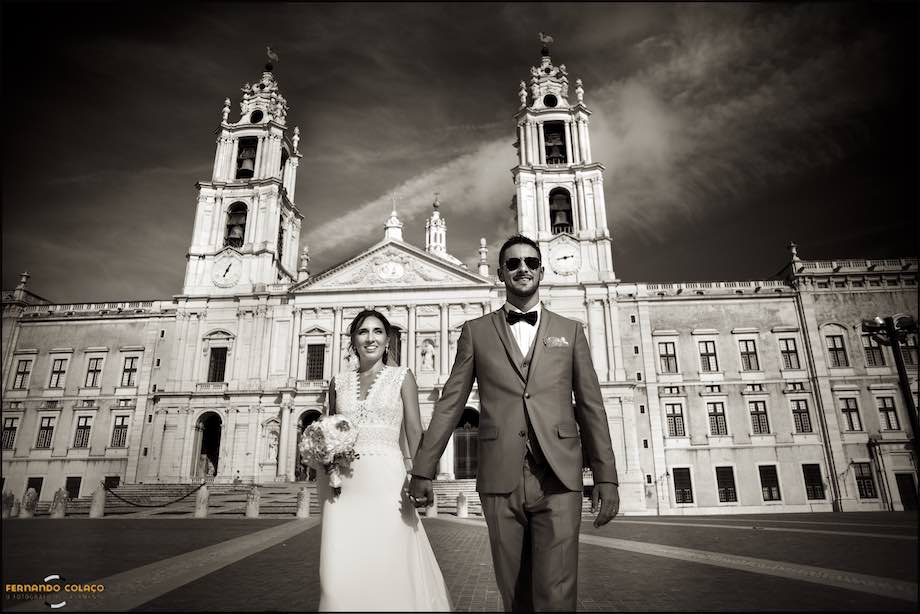 Hand in hand, the bride and groom walk down the sidewalk in front of the Basilica of Mafra on their way to the location of the wedding party.
