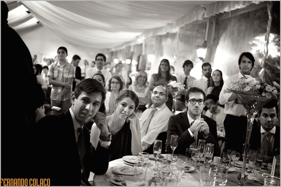 Many wedding guests, some sitting and others standing, look straight ahead at the bride and groom opening up the dance floor.