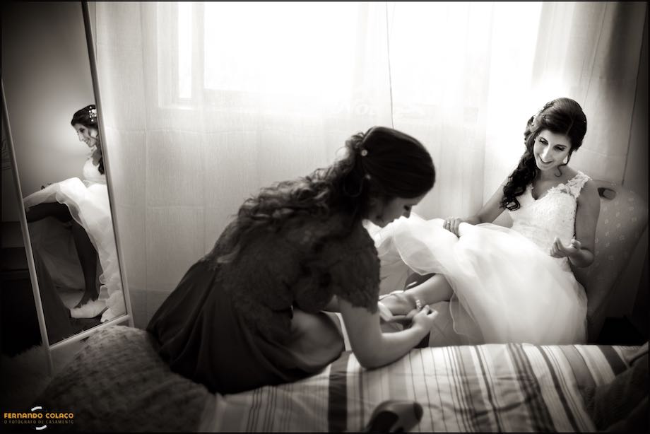 Bride seated and reflected in a mirror, being put on shoes by a friend, in a composition of the wedding photographer in Lisbon.