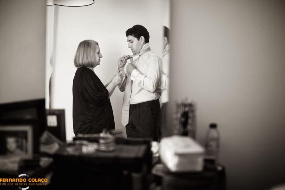 In a mirror, the groom tightening his tie with the help of his mother, seen by the wedding photographer.