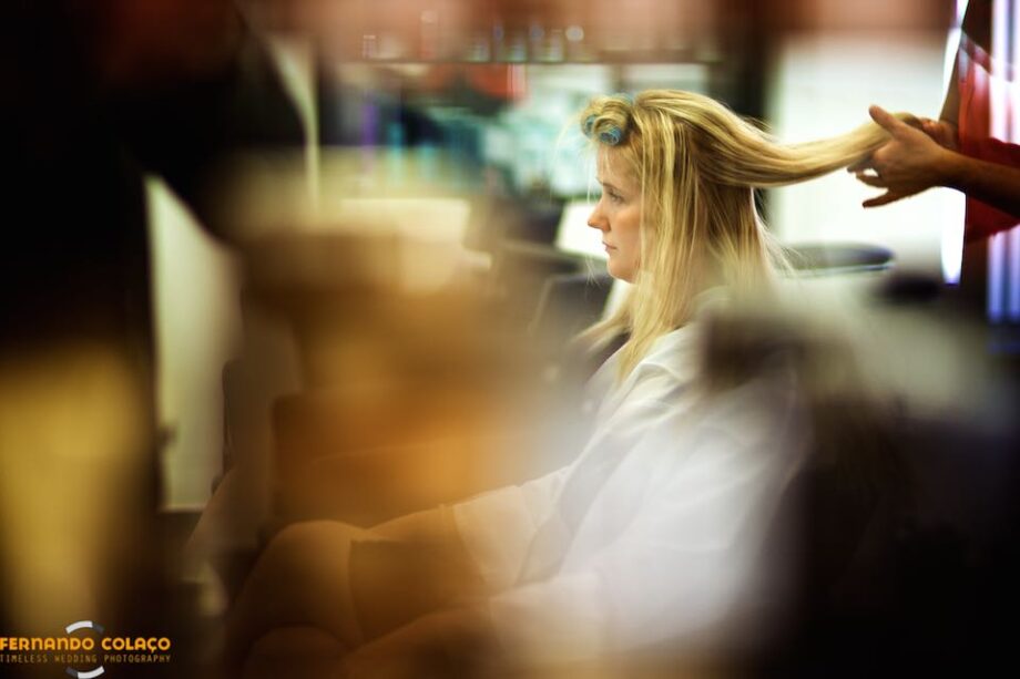 The bride at the hairdresser as her hair is straightened, seen in profile in a mirror by the wedding photographer in Lisbon, Portugal..