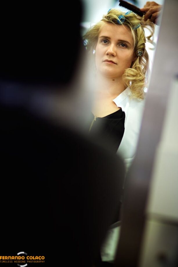In a mirror, the bride seen from the front with the hands of the hairdresser combing her hair.