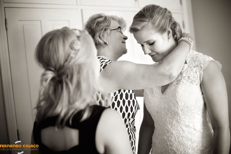 The bride's mother, smiling, puts the necklace around her neck, seen by her sister, from the back.