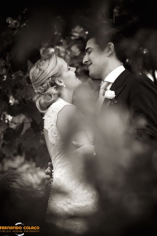 Among the bushes at Quinta dos Alfinetes in Sintra, the bride and groom facing each other smile passionately, seen by the wedding photographer.