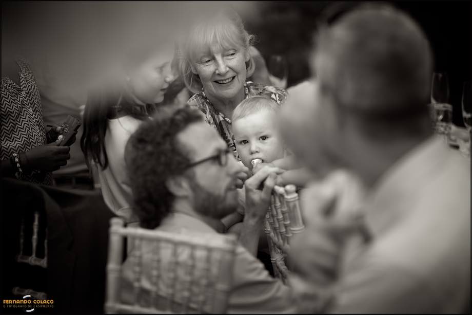 In the middle of other guests sitting at the wedding meal table, a baby on his grandmother's lap.