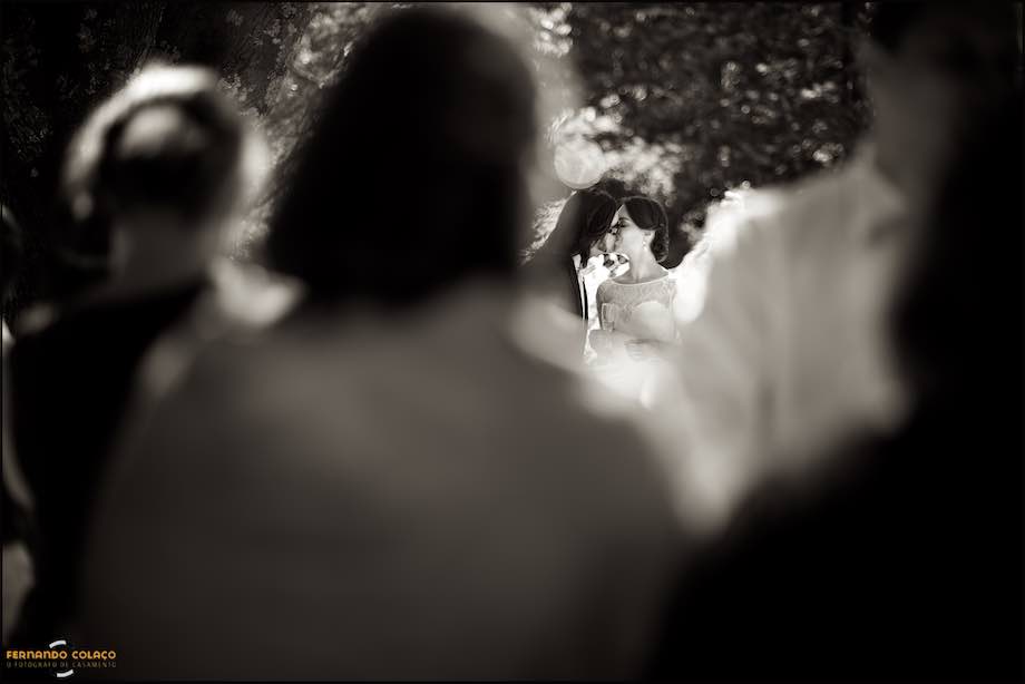 In the distance and among out of focus guests, the bride and groom kiss after the cutting of the wedding cake at Quinta da Serra in Sintra.