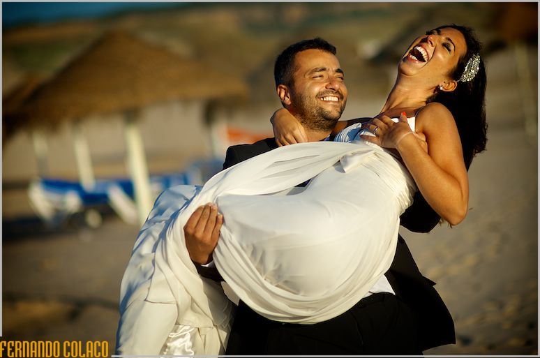 A noiva rindo muito ao colo do noivo que a leva pela praia da Sereia na Costa da Caparica em frente do Waikiki, vistos pelo fotógrafo de casamento em Lisboa.