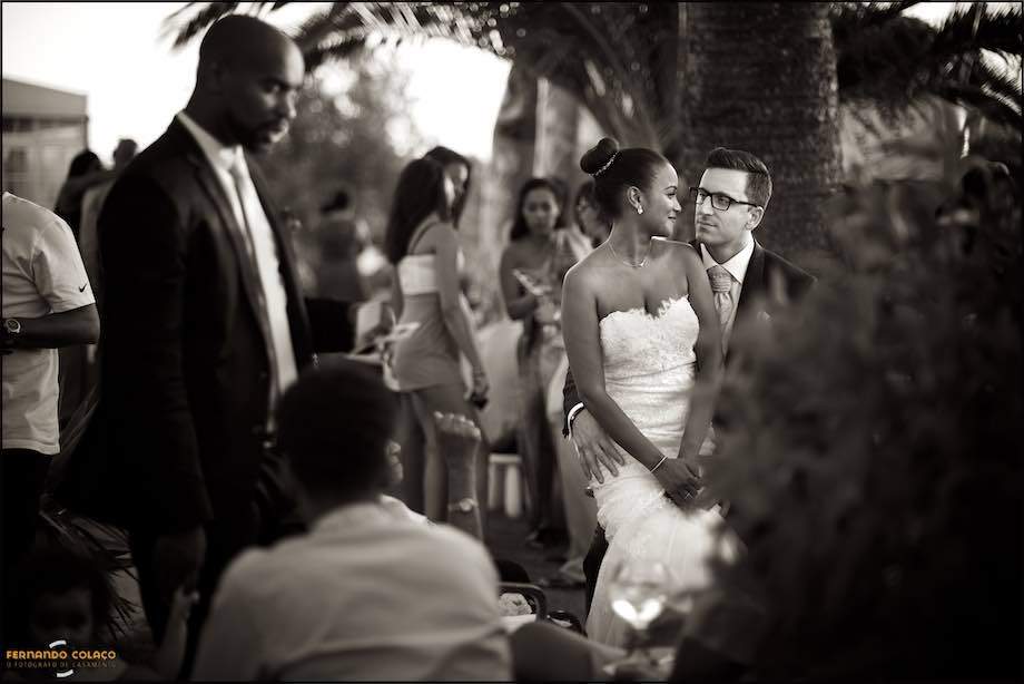Por entre os convidados que convivem no jardim do espaço da festa do casamento e das árvores que lhes fazem sombra, a noiva sentado no colo do noivo e olhando um para outro, conforme, naquele momento, os viu o fotógrafo de casamento em Sintra.