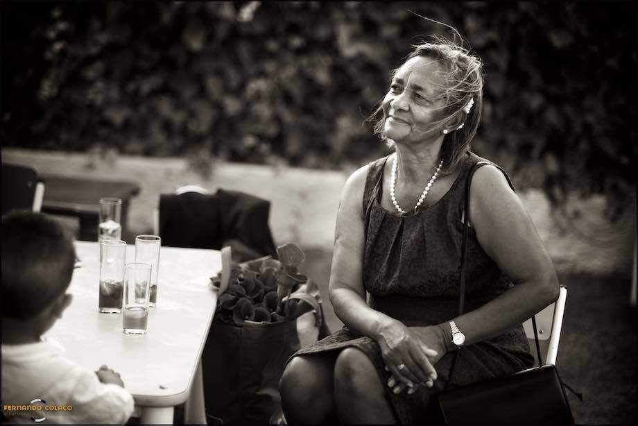 A mãe da noiva, sentada no jardim onde decorre a festa do casamento, olha para o que se passa com um sorriso feliz e satisfeito, assim a viu o fotógrafo de casamento em Sintra.