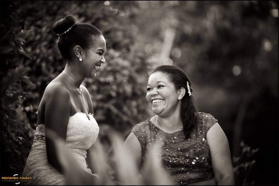 Sentadas sobre uma rocha do jardim do espaço da festa do casamento, a noiva, de perfil e rindo de satisfação, conversa com a sua madrinha que olha para ela sorrindo ternamente, num momento captado pelo fotógrafo de casamento em Sintra.