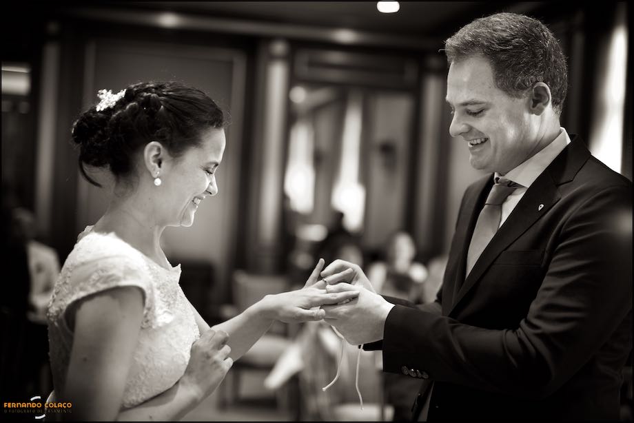 Os noivos, sorridentes, vistos de perfil quando o noivo entrega a aliança do casamento à noiva no Hotel Palácio Estoril.